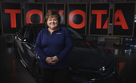 Kim Sweazy, posing for a photograph, wearing a blue Toyota logo polo shirt and smiling in front of a dark-colored Toyota car and the Toyota logo.